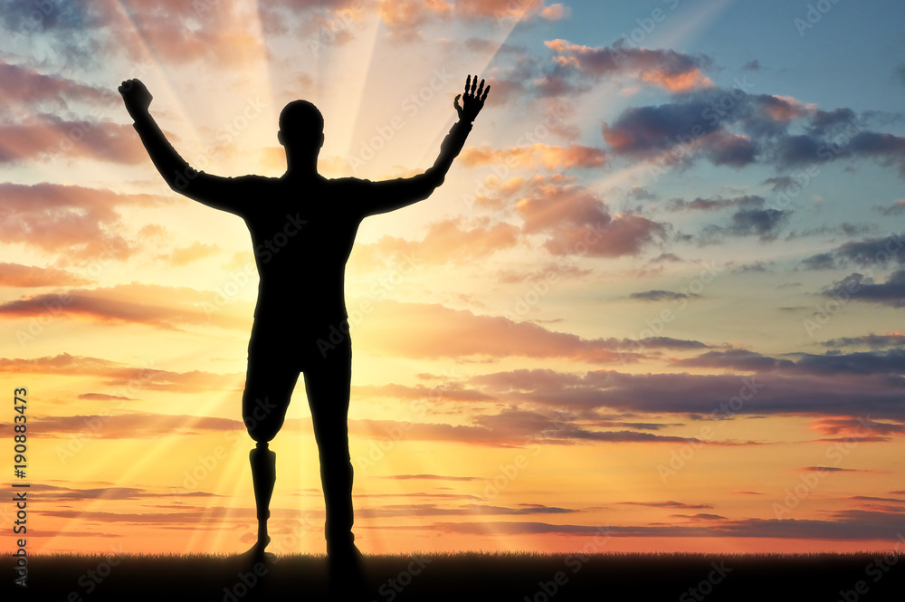 Silhouette of a person with prosthetic leg and arm raising arms in victory at sunrise

